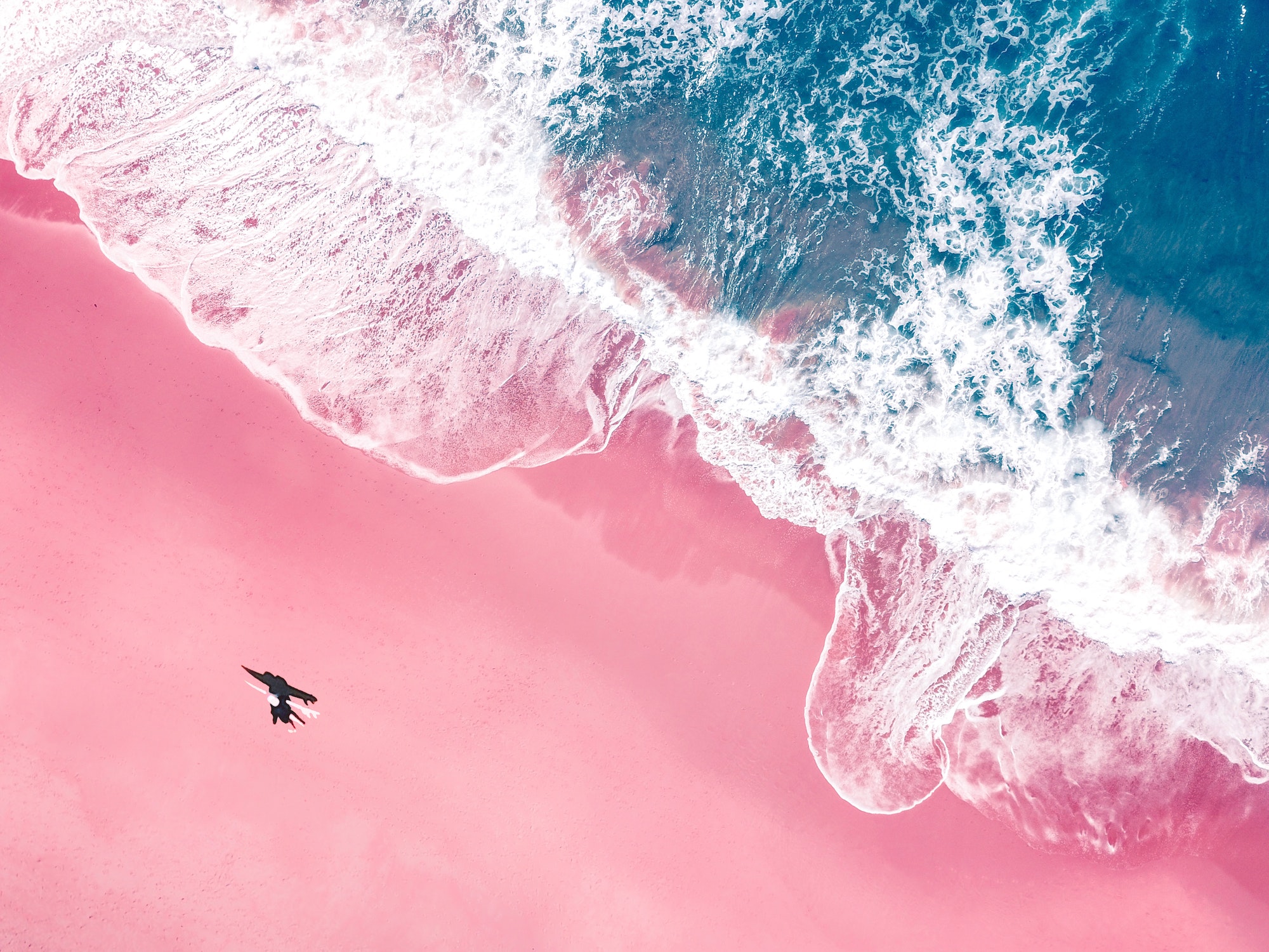 An aerial view of a surfer walking with a surfboard on a pink sandy beach in Australia